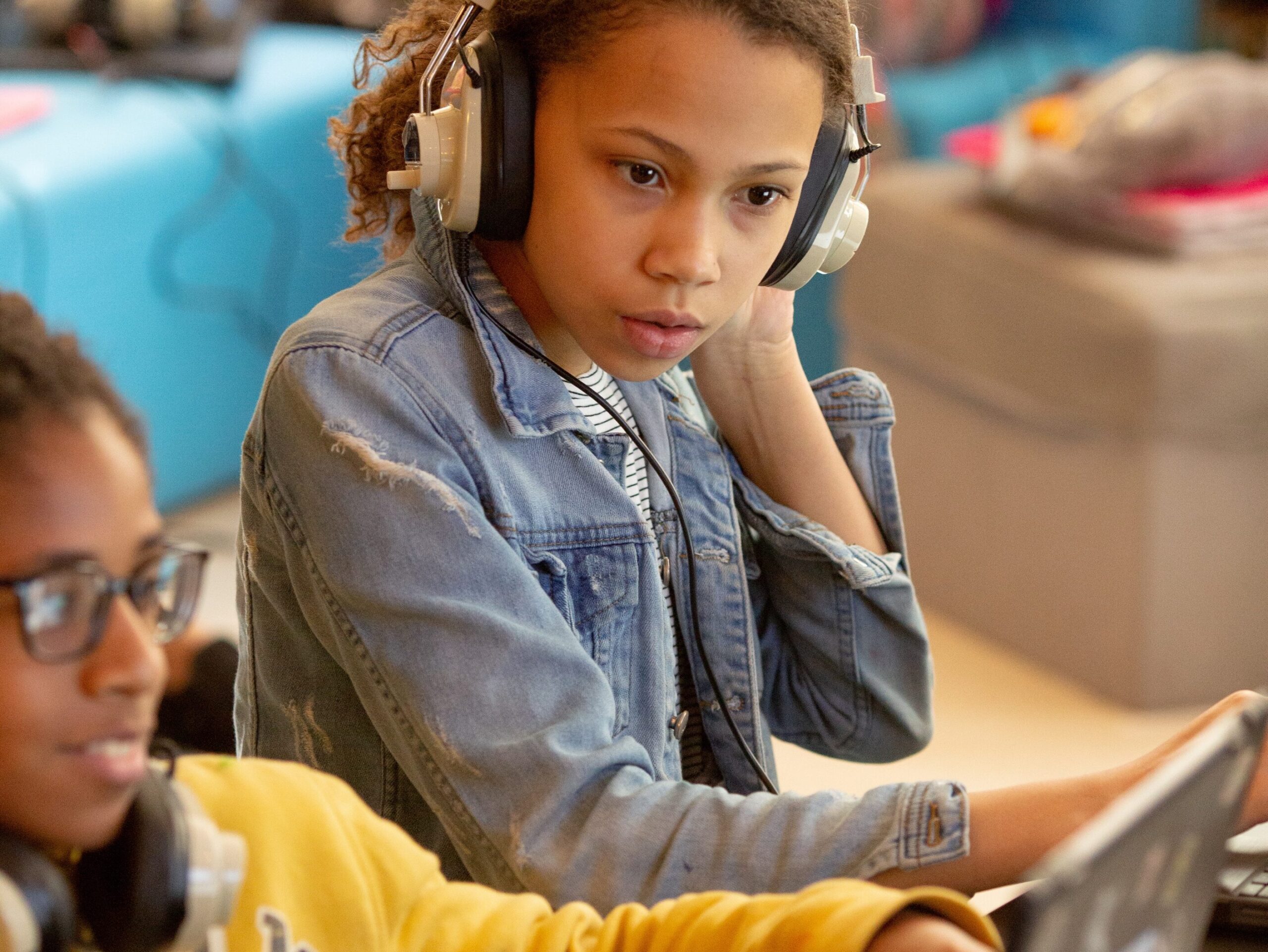 Two students look at a computer, one with headphones on.